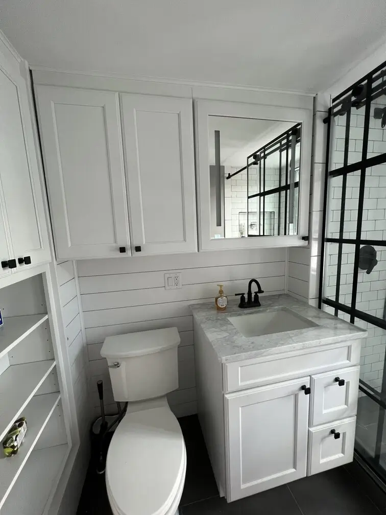 Custom white bathroom vanity with marble countertop, mirrored medicine cabinet, and black-framed glass shower.